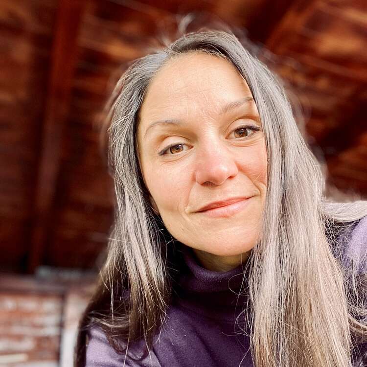 Una mujer con el pelo largo y liso y una sonrisa amable lleva un jersey oscuro de cuello alto. Está en un interior con cálidas vigas de madera de fondo.