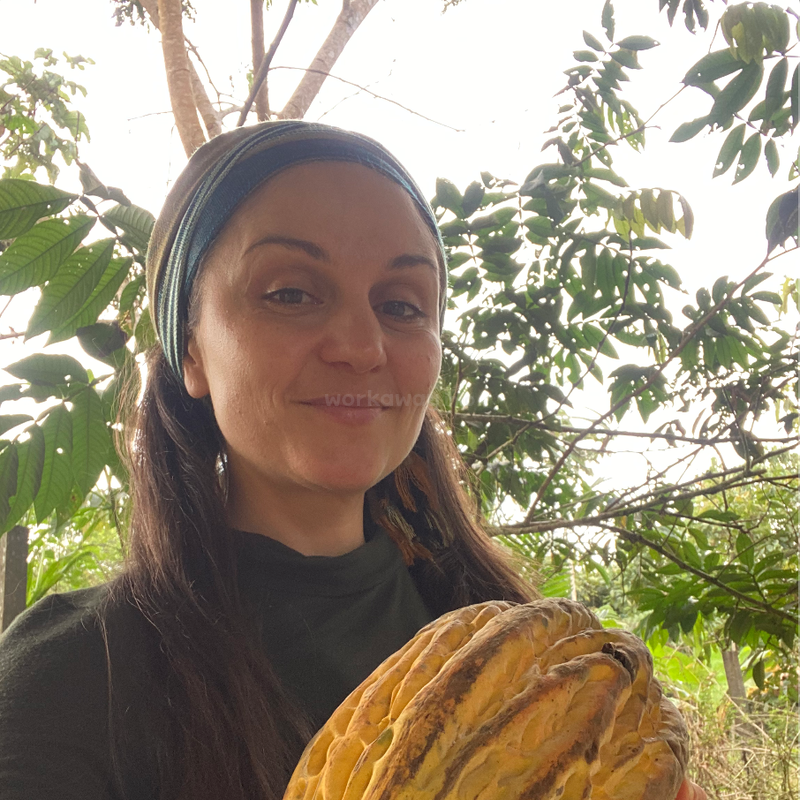 Une femme aux cheveux longs, coiffée d'un bandeau, sourit en plein air, tenant dans ses mains une grosse cabosse de cacao jaune. Des feuilles vertes et des arbres composent le décor naturel en arrière-plan.