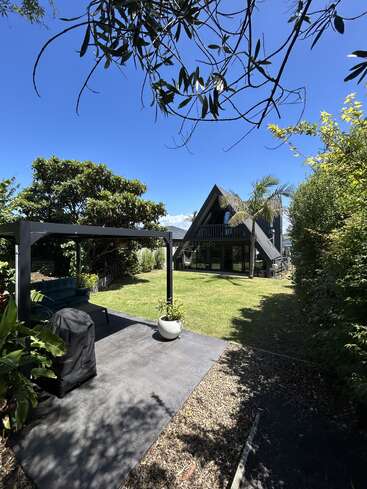 Una moderna casa con techo a dos aguas se encuentra en un jardín soleado y frondoso con palmeras, una zona de descanso exterior a la sombra, vegetación exuberante y un cielo azul brillante.