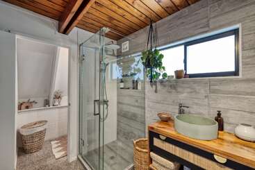 Este cuarto de baño contemporáneo cuenta con una ducha de cristal, techo de madera, lavabo moderno, paredes alicatadas en gris, plantas colgantes, cestas y luz natural procedente de una ventana rectangular esmerilada.