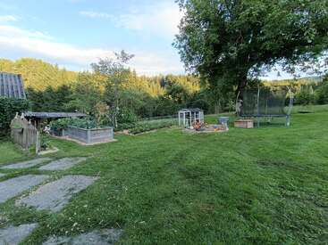 A lush backyard features green grass, raised garden beds, a greenhouse, children’s play area, trampoline, large shade tree, wooden fence, and a peaceful wooded background.