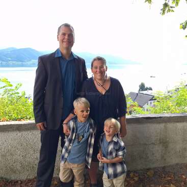 A smiling family of four stands outdoors by a stone wall, with a scenic lake and mountains in the background, dressed in casual and semi-formal attire.
