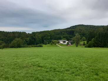 The image depicts a serene landscape featuring a white house situated on a hill, surrounded by lush greenery and a vast grassy field, set against a cloudy sky.