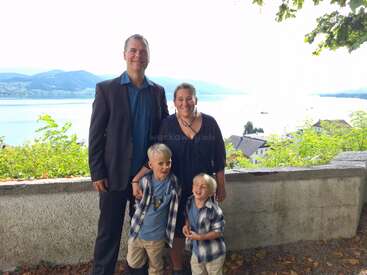 A smiling family of four stands outdoors by a stone wall, with a scenic lake and mountains in the background, dressed in casual and semi-formal attire.