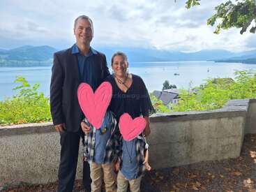 A family of four stands in front of a scenic lake view. The children's faces are covered with pink hearts. The atmosphere is bright and cheerful.