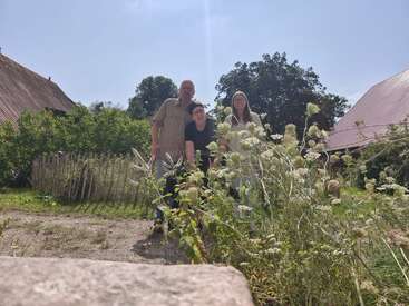 Drei Menschen stehen im Freien im Sonnenlicht und lächeln hinter hohen Wildblumen. Rustikale Gebäude und ein Holzzaun umgeben sie mit Bäumen im Hintergrund; eine entspannte, ländliche Szene.