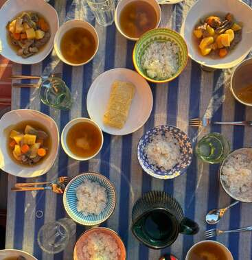 Auf einem bunt gedeckten Tisch stehen Schüsseln mit Reis, Gemüseeintopf, Miso-Suppe, Gläser mit Wasser und ein Teller mit Omelettscheiben auf einer gestreiften Tischdecke.