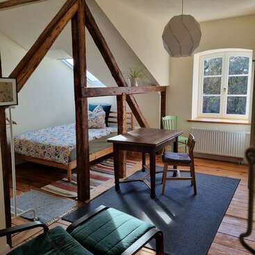 This cozy attic room features exposed wooden beams, a bed with a colorful quilt, a wooden table with chairs, large window, and warm natural sunlight.