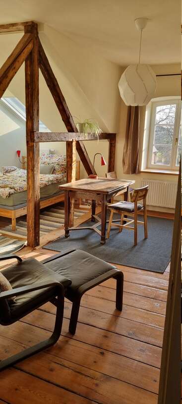Esse quarto aconchegante no sótão tem vigas de madeira, janelas grandes, uma cama com roupa de cama colorida, uma pequena mesa de trabalho, cadeiras, poltrona e luz natural quentinha.