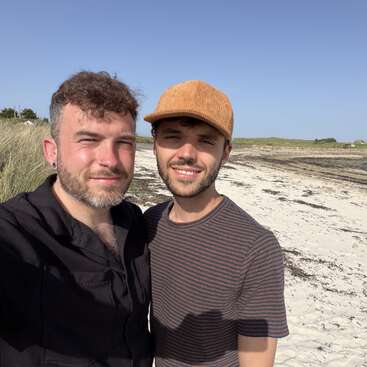 Dos hombres están uno al lado del otro, sonriendo bajo el sol en una playa de arena. Uno lleva una camiseta oscura y el otro, una camiseta a rayas y una gorra de color beige.