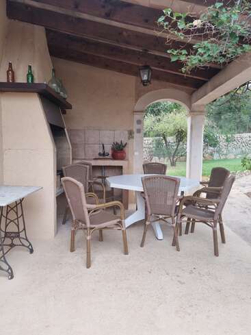 Porche cubierto con techo de madera, zona de barbacoa y fregadero exterior. Seis sillas de mimbre rodean una mesa blanca. Botellas decorativas, una planta en maceta y vegetación en el exterior.