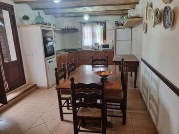 Esta acogedora cocina cuenta con muebles de madera, una mesa central con cuatro sillas, platos decorativos tradicionales en la pared, vigas rústicas y electrodomésticos modernos, como una nevera y un horno.