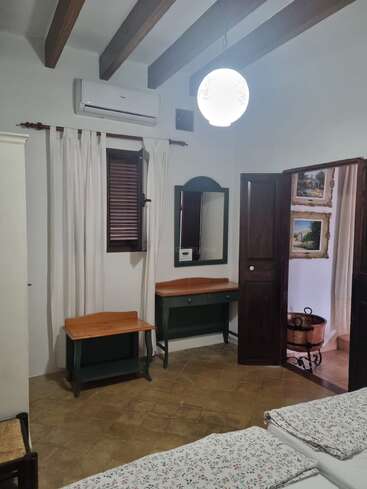 Este acogedor dormitorio cuenta con vigas de madera, aire acondicionado, una lámpara colgante con forma de globo, muebles rústicos, ropa de cama con motivos florales, cuadros en las paredes y una ventana con cortinas blancas.