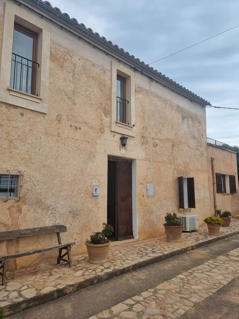 Una casa rústica de piedra de color beige con una puerta de madera, dos ventanas en la parte superior, macetas junto a la entrada, un banco de madera y un cielo nublado sobre ella. Una tranquila escena callejera.