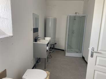 Das ist ein schlichtes, minimalistisch eingerichtetes Badezimmer mit einer Duschkabine, einem Waschbecken mit Spiegel, einem Warmwasserbereiter und einer Toilette. Die Wände und der Boden sind schlicht gehalten.