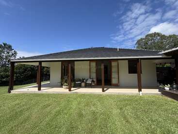 Une maison de plain-pied dotée d'une large véranda, de piliers en bois et de portes vitrées, avec une pelouse soigneusement tondue sous un ciel partiellement nuageux, entourée de verdure et d'arbres.
