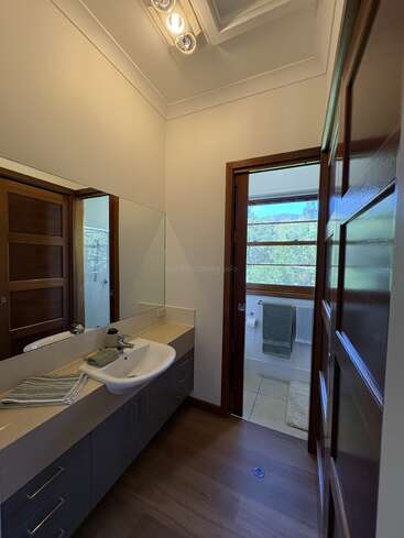 Cette photo montre une salle de bains moderne équipée d'un grand miroir, d'un lavabo, de portes en bois et baignée de lumière naturelle provenant d'une fenêtre. On y aperçoit des serviettes et un tapis de bain.