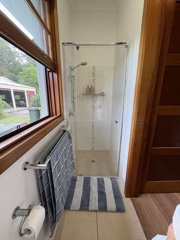 Cette photo montre une salle de bains compacte avec une douche vitrée, un tapis de bain rayé, des serviettes suspendues à un porte-serviettes, un porte-papier hygiénique et une grande fenêtre donnant sur un jardin.