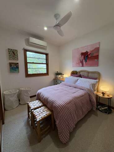 Cette chambre confortable est agrémentée d'un couvre-lit rose à carreaux, de deux tabourets tressés, d'œuvres d'art abstraites, de tables de chevet avec des lampes, d'un climatiseur et d'une fenêtre donnant sur la verdure.