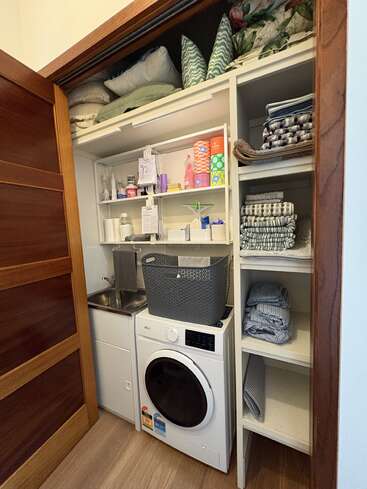 Ce petit local à linge comprend une machine à laver, un évier, des étagères où sont rangés des serviettes soigneusement pliées, du linge de maison, des produits d'entretien et des paniers de rangement. Un espace bien organisé, fonctionnel et parfaitement exploité.
