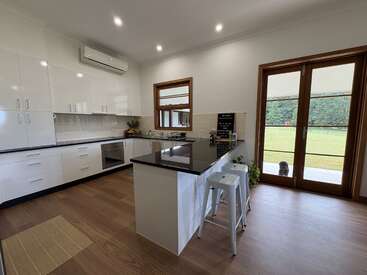 Cette photo montre une cuisine moderne avec des placards blancs, un îlot central au plan de travail noir, des appareils électroménagers en inox, du parquet et de grandes baies vitrées donnant sur l'extérieur. Un chien attend dehors.