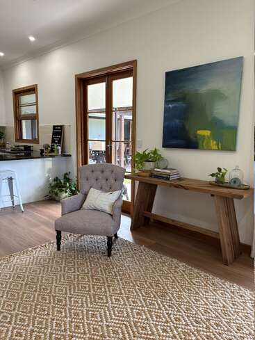Cet espace de vie chaleureux comprend un fauteuil capitonné, un tapis à motifs, une console en bois, des plantes d'intérieur, des œuvres d'art abstrait, de grandes fenêtres et une cuisine attenante baignée de lumière naturelle.