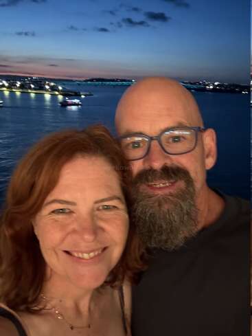 Un couple souriant prend un selfie au coucher du soleil au bord de l'eau. Les lumières de la ville, les bateaux et le littoral pittoresque composent un magnifique décor de soirée romantique.