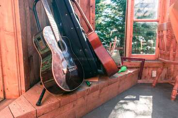 Deux guitares acoustiques sont appuyées contre un mur en bois dans une pièce rustique baignée de soleil. Du matériel musical et des fournitures artistiques sont posés à proximité, et on aperçoit des arbres à travers la fenêtre.