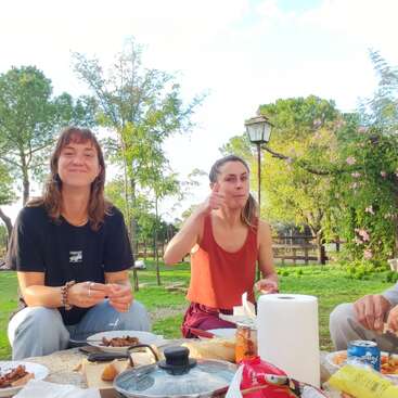 Das Bild zeigt vier Personen, die in einem Park auf dem Boden sitzen, umgeben von einem Picknick-Tisch. Die Gruppe setzt sich aus zwei Frauen und zwei Männern zusammen.