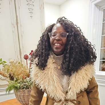 Uma mulher sorridente, com cabelos cacheados e óculos transparentes, usa um casaco de pele aconchegante. Ela está dentro de casa, ao lado de uma cesta de flores secas e folhagens.