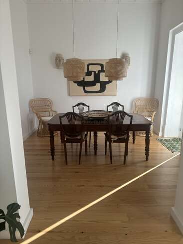 Une salle à manger élégante avec une table en bois, six chaises, des fauteuils en osier, des abat-jours tissés, des œuvres d'art abstraites aux murs et une lumière naturelle sur le parquet.