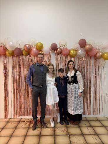 Eine vierköpfige Familie posiert vor einem festlichen Hintergrund mit roségoldenen Fransen und bunten Luftballons. Sie sind schick angezogen und lächeln für das Foto.