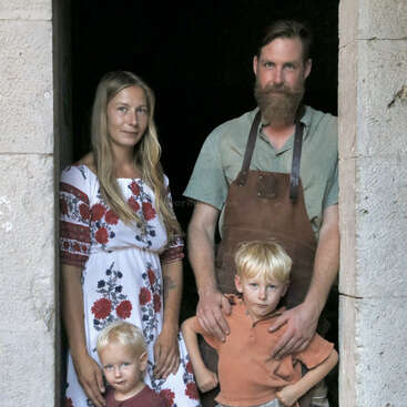 Una familia de cuatro miembros está de pie en el umbral de una puerta: una mujer con un vestido de flores, un hombre barbudo con delantal y sus dos hijos pequeños rubios.