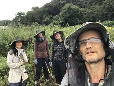 Quatro pessoas usando chapéus, luvas e equipamentos de proteção estão em um cenário verde e exuberante ao ar livre, sorrindo enquanto seguram ferramentas de jardinagem, possivelmente trabalhando em um projeto agrícola.
