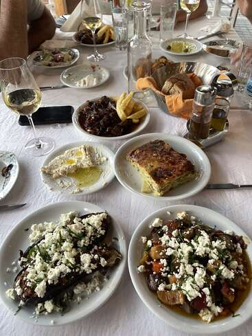 Uma deliciosa refeição mediterrânea servida sobre uma toalha de mesa branca inclui vários pratos: legumes assados com queijo feta, molhos cremosos, pão, vinho e uma suculenta carne acompanhada de batatas fritas. Uma companhia animada.