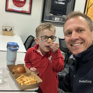 Um homem sorridente e um menino sentam-se à mesa com um hambúrguer e batatas fritas. O menino, de óculos, segura uma batata frita no nariz de forma divertida.