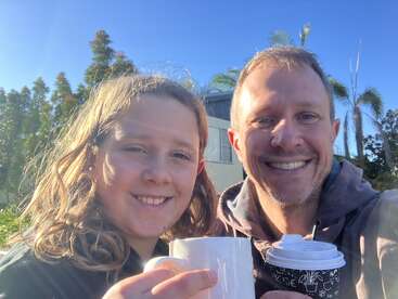 Um homem e uma criança sorridentes estão do lado de fora, segurando xícaras de café. A luz solar intensa, o céu azul e as árvores verdes criam uma atmosfera alegre, relaxada e feliz na foto.