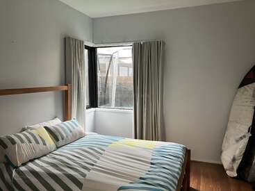 Um quarto pequeno com paredes cinza-claro, cama de madeira, roupa de cama listrada, conjunto de travesseiros, janela grande com cortinas, piso de madeira e uma prancha de surfe no canto.