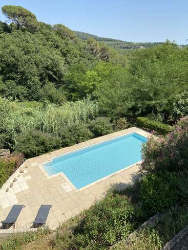 This image shows a rectangular swimming pool surrounded by dense greenery and hills, with two loungers on the tiled poolside, under a clear blue sky.