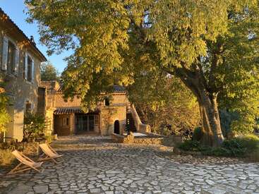 A serene courtyard bathed in golden sunlight features two wooden lounge chairs, rustic stone buildings, a large leafy tree, and a peaceful, inviting atmosphere.