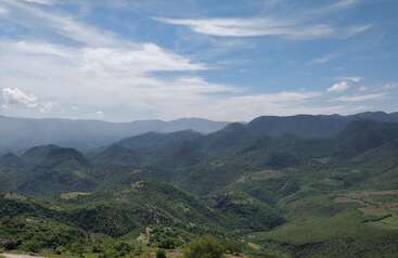 In der Ferne erstrecken sich grüne Berge unter einem strahlend blauen Himmel, der mit Wolkenfetzen übersät ist. Das Sonnenlicht beleuchtet die üppige Landschaft und schafft eine friedliche, ruhige Szene.