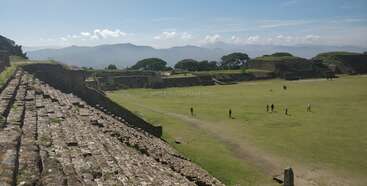 Alte Steinruinen mit Stufenstrukturen umgeben ein weites Grasfeld. In der Ferne rahmen Berge und Bäume die Szene ein, während kleine Gruppen von Menschen das Gelände erkunden.
