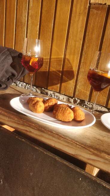 Zwei Gläser mit rotem Getränk und Orangenscheiben stehen neben einem weißen Teller mit fünf goldbraun gebratenen Snacks auf einem rustikalen Holztisch.