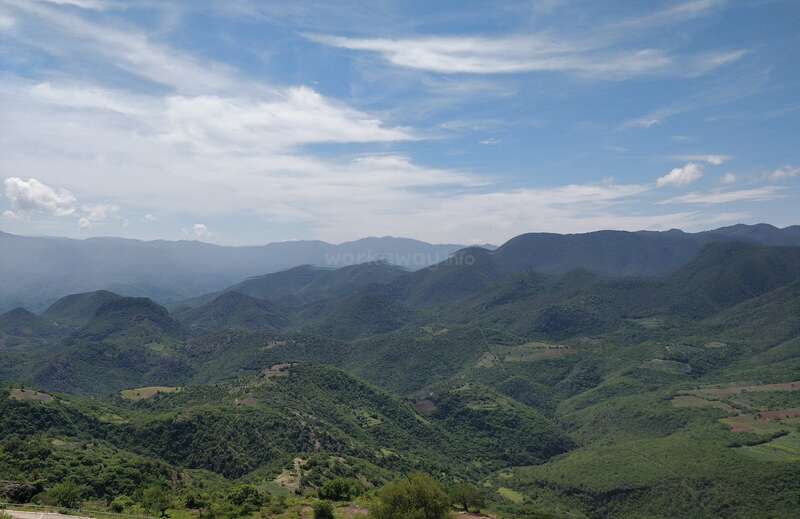 In der Ferne erstrecken sich grüne Berge unter einem strahlend blauen Himmel, der mit Wolkenfetzen übersät ist. Das Sonnenlicht beleuchtet die üppige Landschaft und schafft eine friedliche, ruhige Szene.