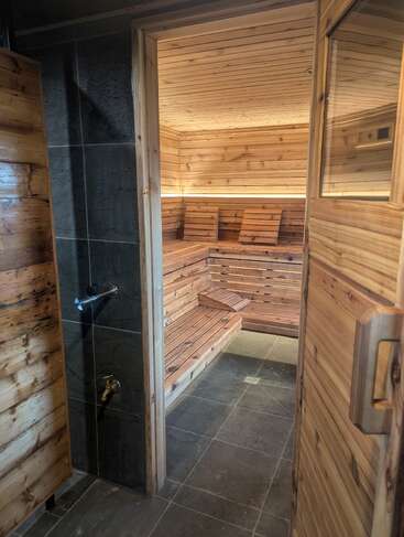 Esta imagen muestra una moderna sauna de madera con bancos y respaldos. La puerta está abierta, dejando ver el suelo de baldosas y la cálida iluminación ambiental del interior. Un ambiente acogedor.