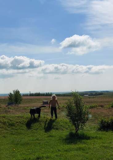A young child with long hair runs through a field with a black dog, surrounded by trees and a blue sky with white clouds.