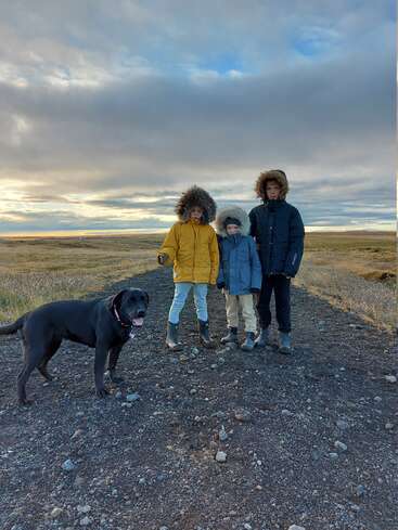 Three children and a dog stand on a dirt path in a vast, grassy field under a cloudy sky, all wearing warm winter coats.