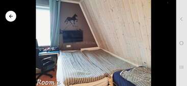 A cozy attic room with slanted wooden walls, twin beds, striped bedding, wall art of a horse, desk with chair, window with curtains, and television.