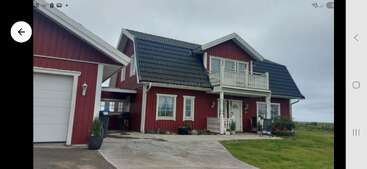 A charming red two-story house with a black roof, white trim, front porch, balcony, attached garage, paved driveway, and surrounded by green grass under a cloudy sky.