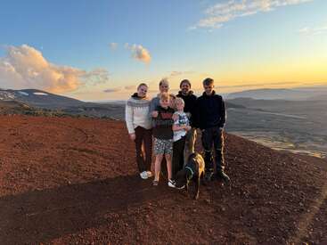 A family of five and their dog stand together on a rocky hilltop at sunset, surrounded by a scenic landscape of mountains and dramatic clouds.